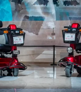 Two red mobility scooters are parked indoors on a shiny floor. Each scooter has a front basket with attached signage. A blurred mural is visible in the background.