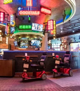 Two red mobility scooters parked indoors under colorful neon signs and multiple TV screens in a lively bar or entertainment venue.