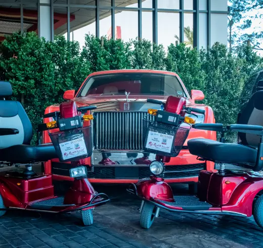A red Rolls-Royce car parked behind two red mobility scooters with contest signs attached to their handlebars. Trees and a building are in the background.