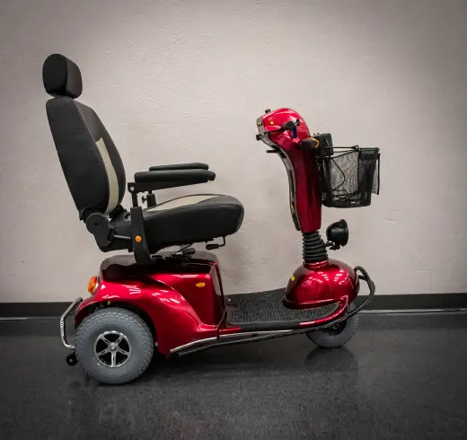 A red mobility scooter with a black seat and small front basket is positioned against a neutral wall.