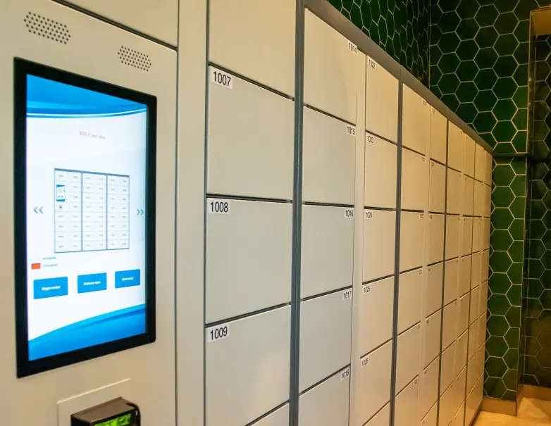 A row of numbered lockers with a digital touchscreen interface on the left side for accessing and managing lockers. The wall has a green hexagonal tile pattern.